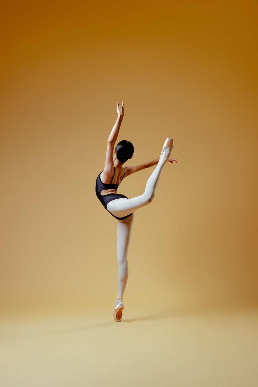 A ballet dancer in a black leotard balancing en pointe with one leg lifted behind her in a studio portrait.