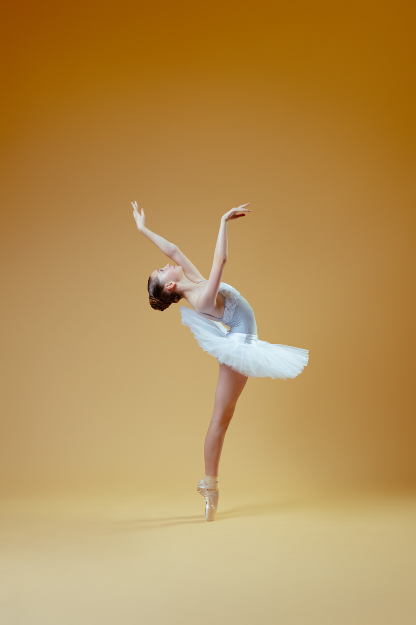 A ballet dancer in a white tutu arching backward with her arms raised in a studio portrait.